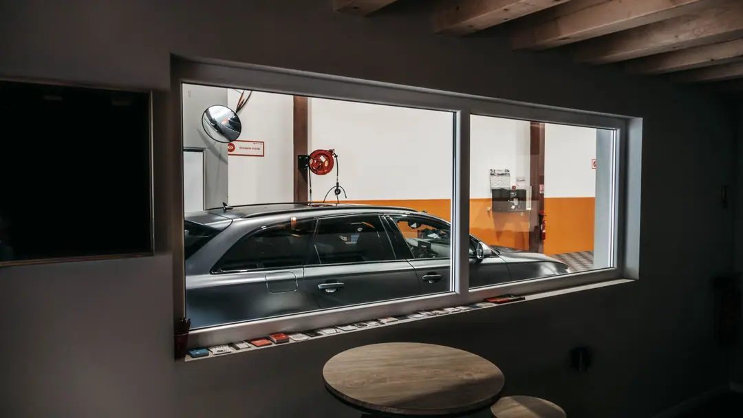 Salle d'attente sombre donnant par une large baie vitrée sur un break gris stationné dans une ligne de contrôle technique